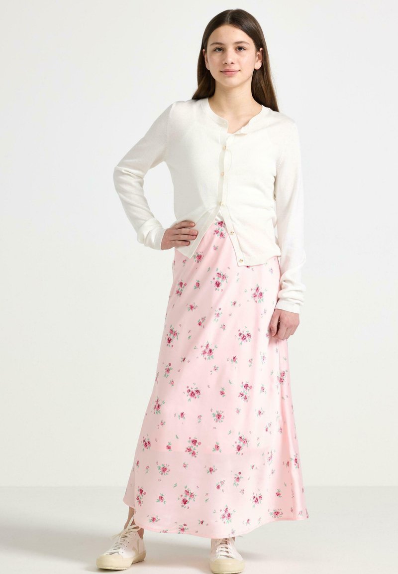 Witte lange mouwen cardigan met knopen over een roze bloemen maxi-jurk met kleine rozen en groene bladeren, gecombineerd met witte sneakers.