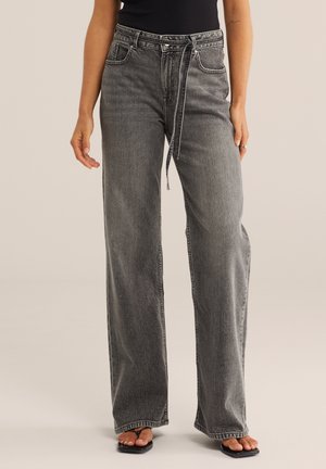 Naine kannab kõrge vöökohaga, laiade säärtega halli teksapükse koos sobiva kangast vöö, musta toppi ja musti varbavahe sandaale.