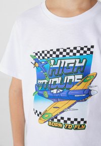 Biała koszulka z kolorową grafiką samolotów oraz napisem "WYSOKA POSTAWA" i wzorem w kratkę na górze.