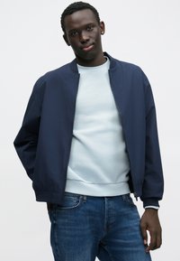 Marineblauwe bomberjack met een ronde kraag, ritssluiting en lichtgewicht stof. Draagt over een lichtblauwe trui en spijkerbroek.