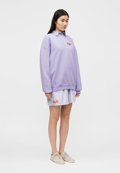 Γυναίκα με μακριά σκούρα μαλλιά που φοράει ένα υπερμεγέθες φουτεράκι λεβαντίνης πάνω από ένα μπλε καρό φόρεμα και μπεζ αθλητικά παπούτσια, στέκεται σε απλό φόντο.