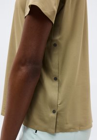 Δεξί χέρι ατόμου που φοράει υφασμάτινο, κοντομάνικο μπεζ πουκάμισο με μαύρα κουμπιά τύπου snap κατά μήκος της πλαϊνής ραφής.