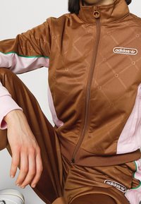 Person wearing brown Adidas tracksuit with pink and green accents, sitting with one leg bent and hand resting on the knee.