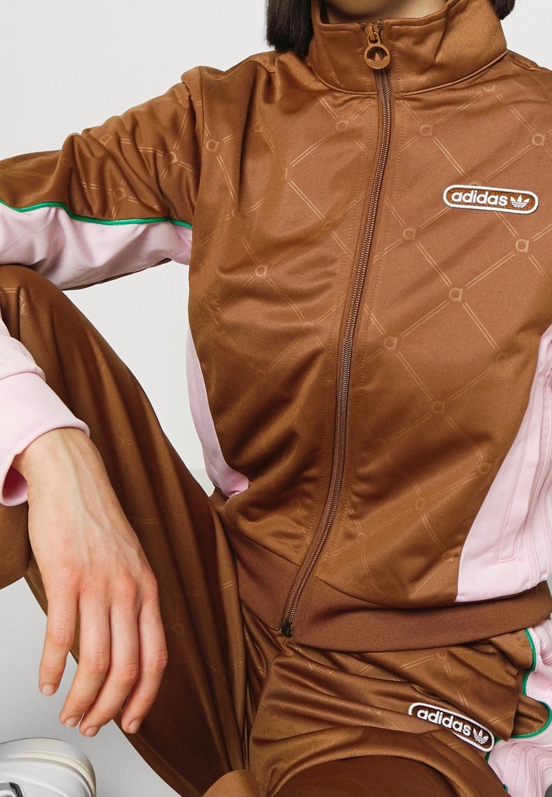 Person wearing brown Adidas tracksuit with pink and green accents, sitting with one leg bent and hand resting on the knee.