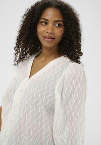 Femme aux cheveux bouclés portant une blouse blanche texturée avec des boutons, regardant légèrement sur le côté devant un fond neutre.