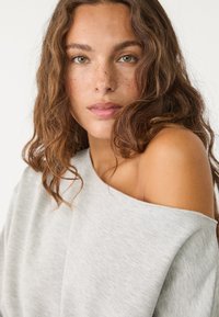 Helles graues Off-Shoulder-Oberteil aus weichem Stoff, mit einer lockeren Passform und minimaler Textur. Akzentuierung des natürlichen Hauttons und welligem Haar.