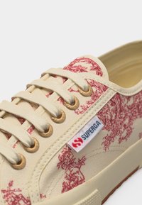 Nærbilde av beige lerretssneakers med rødt blomsterprint, beige lisser gjennom gullfargede maljer og Superga-logo på siden.
