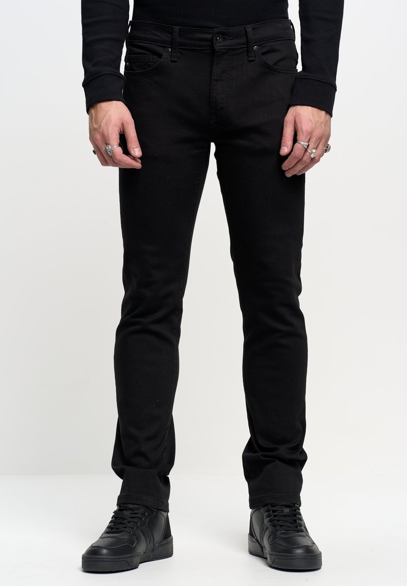Jeans neri slim-fit con cinque tasche, realizzati in denim elasticizzato, caratterizzati da una texture liscia e dettagli di cucitura sottili, abbinati a sneakers nere.