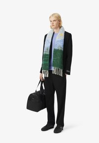 Blonde persoon staat met een zwarte sporttas, gekleed in een zwart jasje, lichtblauwe shirt en een kleurrijke sjaal met een landschapdesign.