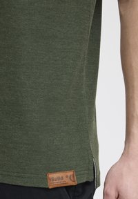Olivengrøn bomulds t-shirt med en struktureret overflade, sideslids og et brunt mærke i læder ved kanten.
