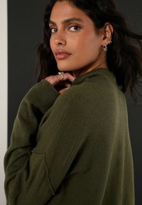 Sweter oliwkowy, miękki materiał, luźny krój, ściągacze przy mankietach i dekolcie. Modelka pozuje z ręką blisko twarzy, prezentując kolczyki w kształcie kół.