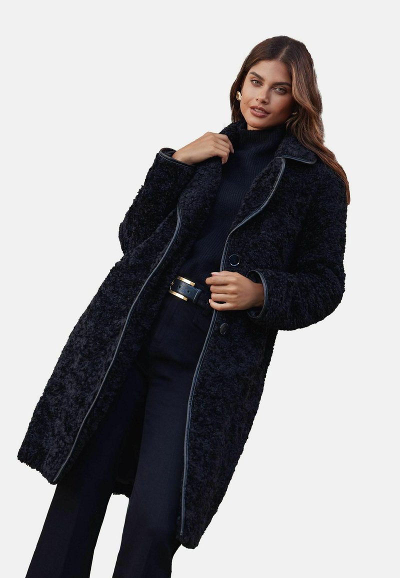 Palton lung, negru, texturat, dintr-un material plușat, cu chenare contrastante și nasturi mari; purtătorul ține gulerul, asortat cu un helancă neagră.