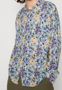 Camisa de botones de manga corta en azul claro con un patrón tropical que presenta tonos terrenales, palmeras y diseños abstractos.