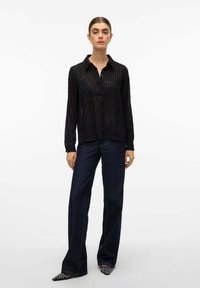 Camicia nera trasparente con bottoni, a righe verticali, adornata da piccole paillettes; abbinata a jeans a gamba larga blu scuro e scarpe con punta.