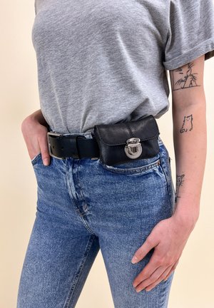 Svart läderpung med ett silverlås, bärs på en blå denimmidja. Modellen har på sig en grå t-shirt och har tatueringar på armen.