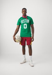 Grøn basketballtrøje med "BOSTON" og nummer "0," røde shorts med gule striber, hvide sneakers og en basketball i hånden.