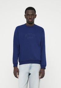 Sweatshirt bleu marine à col rond, avec des poignets et un ourlet côtelés. Présente un texte brodé "ORIGINALS Studio" à l'avant. Matière : coton.