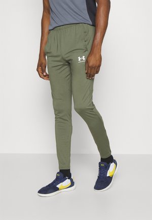 Mand iført olivengrønne Under Armour joggebukser og marineblå sneakers med gul swoosh, stående mod en ensfarvet lys baggrund.