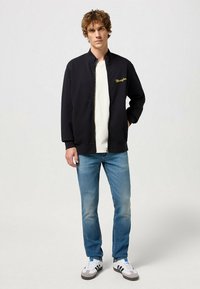 Chaqueta negra con cierre y puños acanalados, que presenta un bordado dorado de "Wrangler" en el pecho, llevada sobre una camiseta blanca y jeans azules.