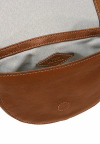 Binnenkant van een bruine leren tas met een gestructureerde grijze voering, voorzien van een gestikte Fossil-logo tag en een circulaire accent aan de onderkant.