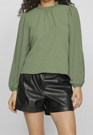 Kvinde iført en olivengrøn bluse med lange ærmer og flæser ved kraven samt sorte shorts i imiteret læder mod en ensfarvet baggrund.