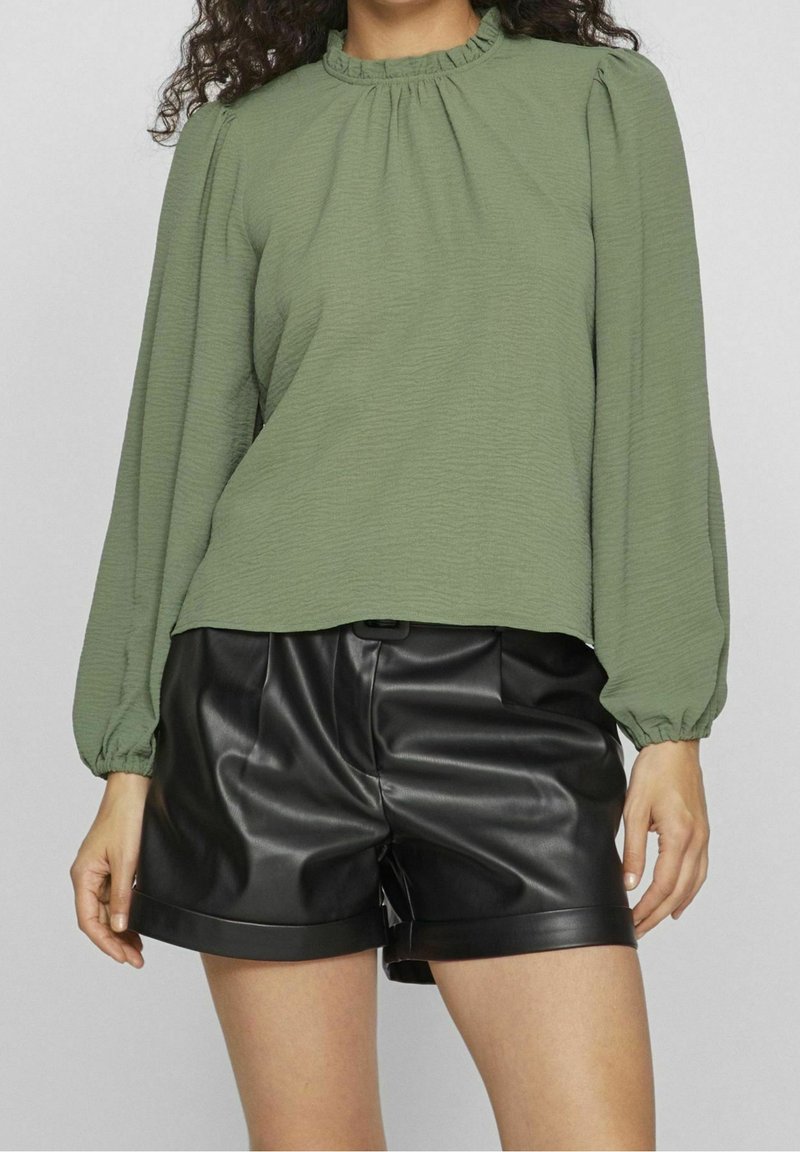 Femme portant un chemisier vert olive à manches longues avec un col à volants et un short noir en simili-cuir sur fond uni.