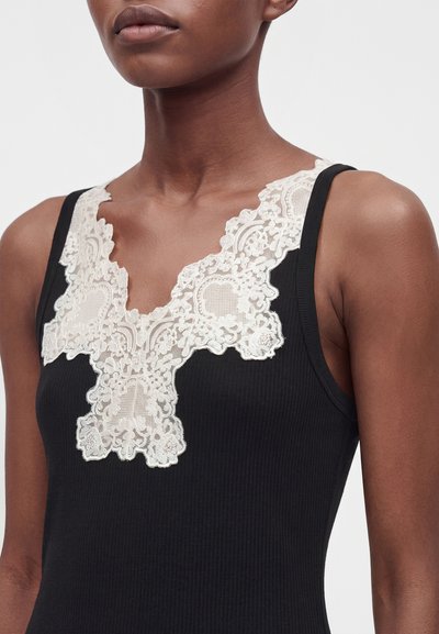 Femme portant un haut noir sans manches avec un décolleté en V profond orné de dentelle florale blanche.