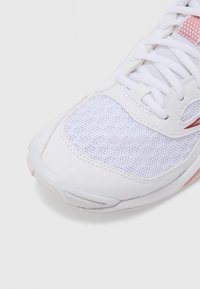 Valkoinen urheilukenkä, jossa on verkkokangasta ja synteettisiä materiaaleja, pyöreä kärki, nauhat ja hienovarainen punainen korostus lähellä pohjaa.