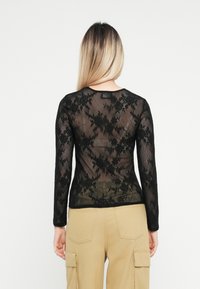 Top lungo nero a maniche con pizzo a motivi floreali, caratterizzato da un collo rotondo e una trama semi-trasparente. Indossato con pantaloni cargo beige.