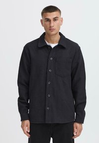Black, textured shirt jacket with a collar, button closure, and front pocket. Features a ribbed pattern and long sleeves.