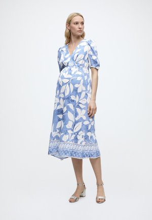 Raskaana oleva nainen, joka pukeutunut sinivalkoiseen kukalliseen midimekkoon, jossa on pussihihat, ja hopeanväriset, paksukorkoiset sandaalit, seisomassa valkoisella taustalla.