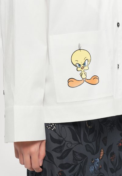 White shirt pocket with angry yellow cartoon bird illustration, worn with dark floral patterned skirt, hand relaxed at side.