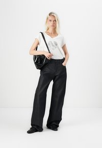 T-shirt blanc avec un graphic de cœur, associé à un jean large bleu foncé. Sac bandoulière noir. Le mannequin se tient contre un fond blanc uni.