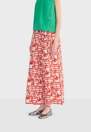 Osoba v širokých nohaviciach s červeným abstraktným vzorom, zelenom tielku bez rukávov a metalických strieborných balerínkach, stojaca pred jednoduchým pozadím.