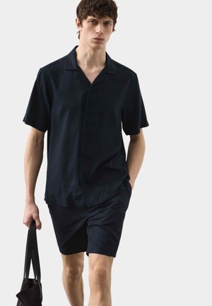 Młody mężczyzna idzie, ubrany w czarną koszulkę z krótkim rękawem i spodenki, trzymając w lewej ręce czarną torbę na ramię.