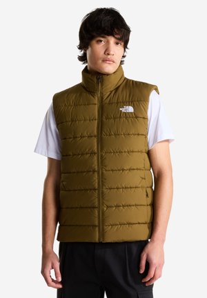 Giovane uomo indossa un gilet trapuntato verde oliva con cerniera sopra una camicia bianca a maniche corte, in piedi davanti a uno sfondo semplice.