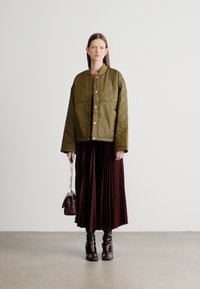 Pihetszerű, olívazöld bomberdzseki kerek nyakkivágással, hosszú, pliszírozott bordó szoknyával és térdig érő sötétbarna csizmával párosítva.