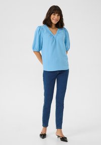 Light blue, short-sleeve blouse with a V-neck and gathered details; paired with dark blue skinny jeans and black heeled mules.