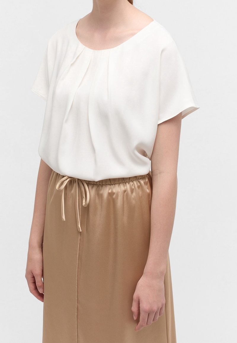 Vit kortärmad topp med veck, kombinerad med en ljus beige satinskjol med elastisk midja och dragsko-detalj.