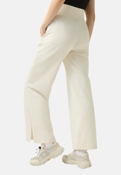 Wide-leg cream pants with a smooth texture, side pockets, and a high waistband; features a split detail at the hem. Paired with light sneakers.