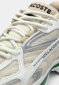 Gros plan d'une basket Lacoste beige et blanche avec des lacets blancs, des accents argentés et du vert sur la semelle. Le logo Lacoste est visible sur la languette et le côté.