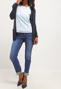 Cardigan marine, haut bleu clair et jeans bleus à revers. Des talons sombres soulignent la tenue. Tissu lisse, design ajusté et formes classiques.