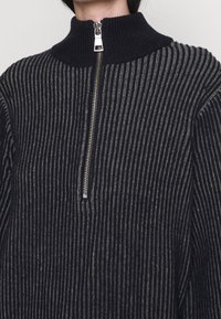 Nahaufnahme einer Person, die einen schwarz-grau gestreiften Strickpullover mit einem silbernen Halbreißverschluss und einem gerippten Stehkragen trägt.