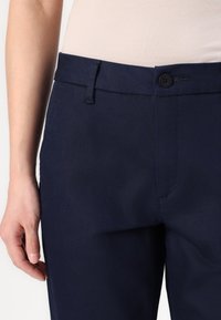 Granatowe spodnie o gładkiej fakturze, z płaskim przodem, zapięciem na pojedynczy guzik oraz subtelnym detalem bocznych kieszeni.
