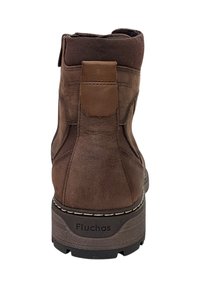 Botín de cuero marrón con un acabado texturizado, paneles laterales elásticos y una suela de goma con tachuelas. Logo "Fluchos" embossado en el talón.
