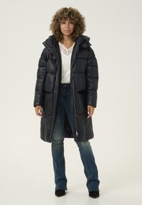 Abrigo acolchado negro con capucha, con cierre delantero, textura acolchada y larga longitud. Lleva puesto un top blanco y unos vaqueros azules.