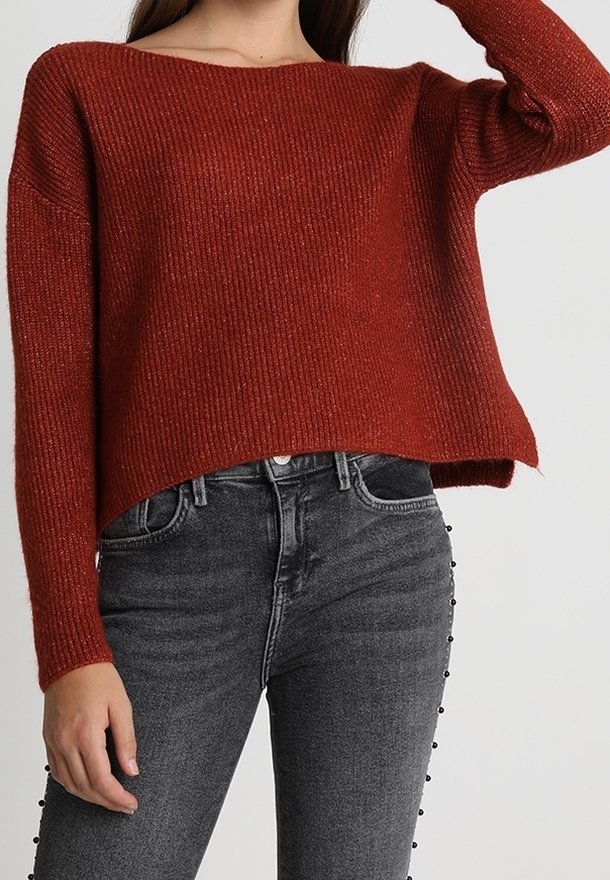 Rostfarbener, gerippter Strickpullover mit weitem Ausschnitt und langen Ärmeln; kombiniert mit hochgeschnittenen, dunkelgrauen Jeans mit Knopfdetails.