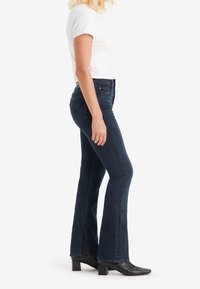 Pantalones de mezclilla acampanados de color azul oscuro, con una cintura alta, aberturas laterales y un silueta ajustada, combinados con botas de tobillo negras con tacón.