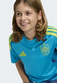 Ljusblå sporttröja med gula accenter på axlarna, som har Spaniens landslagsemblem och tre-randig design.