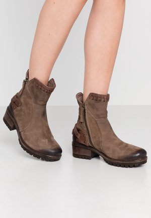 Bottes en cuir marron montant à la cheville, avec fermetures éclair latérales, lanières décoratives et talons épais, portées sur des jambes nues sur un fond uni.
